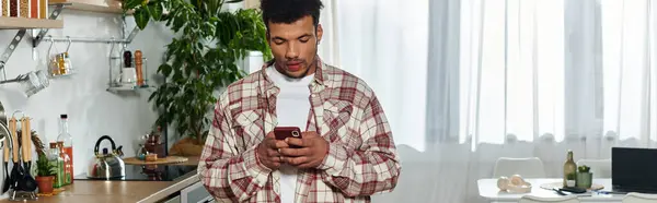 Handsome young man stands in a stylish kitchen using his smartphone and looking focused — Stock Photo