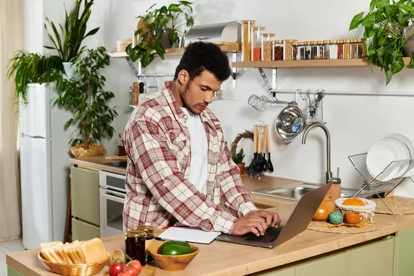 A young man focuses on his laptop while standing in a stylish kitchen surrounded by fresh food. — Stock Photo