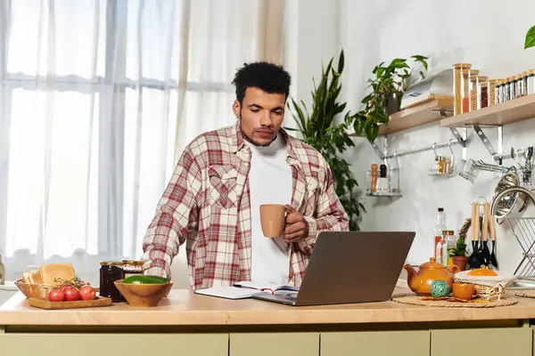 A young handsome man is sipping coffee while focused on his laptop in a bright kitchen. — Stock Photo