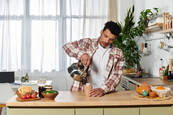 Un joven guapo vierte agua caliente en una taza, preparando té en una cocina elegante. - foto de stock