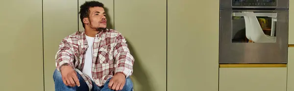 A young handsome man is thoughtfully sitting on the floor in a contemporary kitchen setting. — Stock Photo
