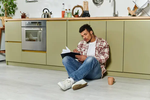 Handsome young man relaxes on the floor in a modern kitchen while enjoying a book and coffee. — Stock Photo