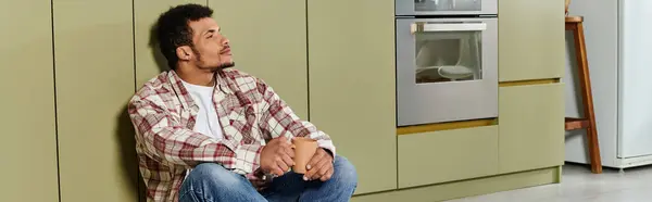 Handsome young man relaxes on the floor of a stylish kitchen while holding a warm drink. — Stock Photo