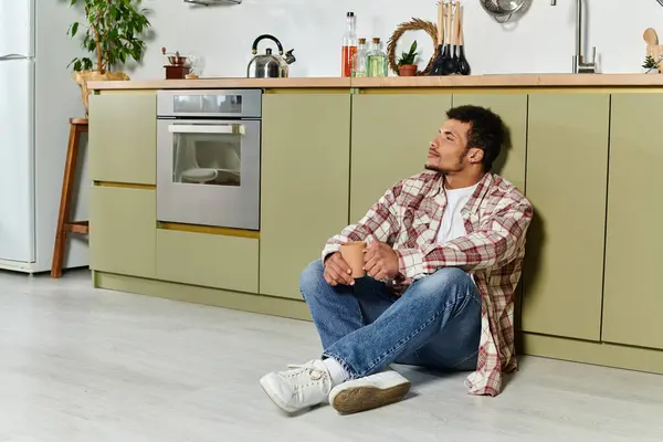 A young handsome man relaxes on the floor of a stylish kitchen while holding a cup. — Stock Photo