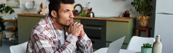 Thoughtful young man dressed casually sits at a kitchen table using a laptop and reflecting. — Stock Photo