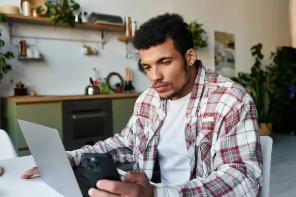 Un jeune homme beau se concentre sur son smartphone tout en travaillant sur un ordinateur portable à la maison. — Photo de stock