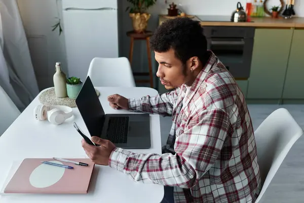 Young man focuses on his laptop while holding his phone in a stylish kitchen setting. — Stock Photo