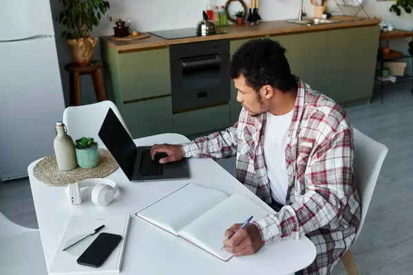 Young man focused on his laptop while taking notes in a cozy kitchen setting. — Stock Photo
