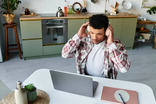 A young man listens to music on headphones while focusing on a laptop in a modern kitchen. — Stock Photo