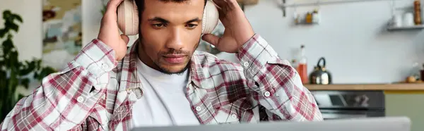 Guapo joven disfruta de la música a través de auriculares mientras está comprometido con su computadora portátil en casa. - foto de stock