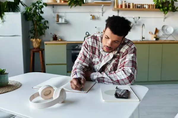A young handsome man writes thoughtfully in his notebook while sitting at a stylish kitchen table. — Stock Photo