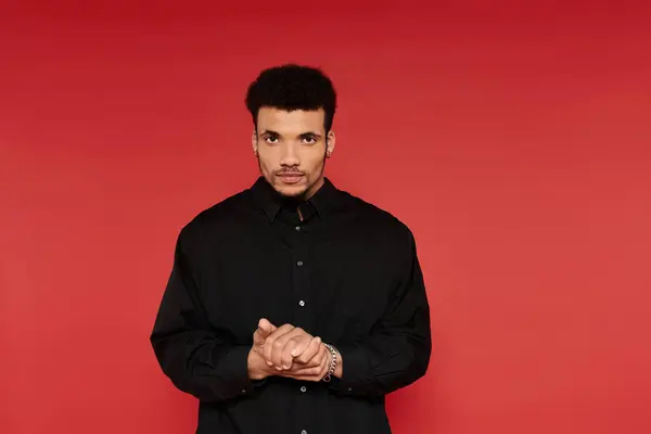 Un joven guapo se para con las manos apretadas, con una camisa negra, frente a un telón de fondo rojo. - foto de stock