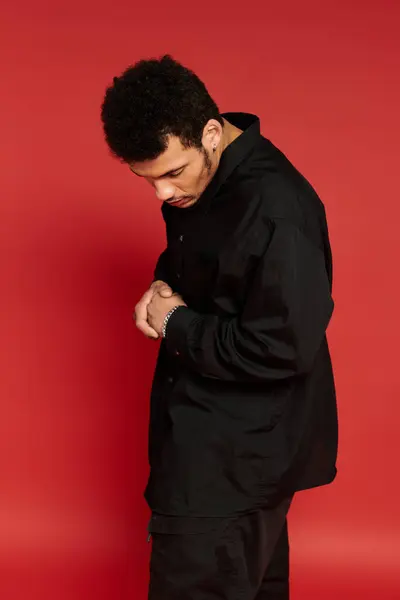 Young man looks down thoughtfully while dressed in a black shirt, set against a bright red backdrop. — Stock Photo