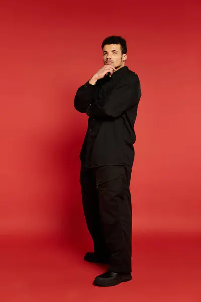 Young man stands confidently with one hand on his chin, wearing black attire on red backdrop. — Stock Photo