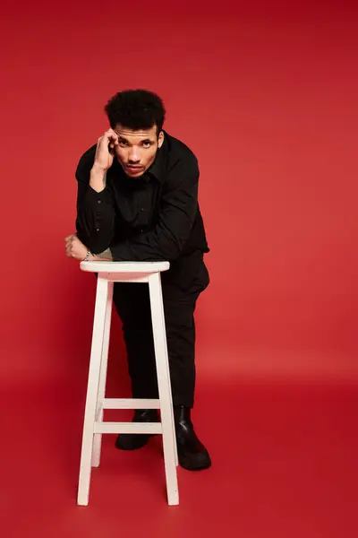 A young handsome man leans on a stool, posing thoughtfully against a bold red backdrop. — Stock Photo