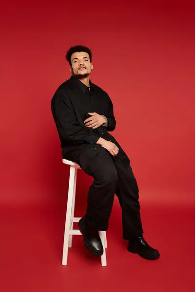 Young man poses confidently on a white chair in front of a bold red backdrop. — Stock Photo