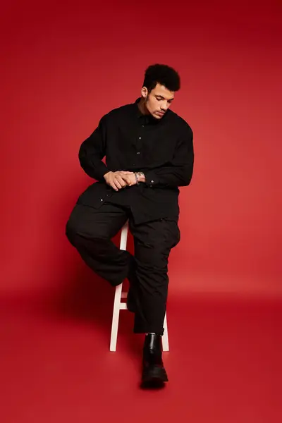 A young man sits on a stool, wearing a black outfit and looking thoughtful in a bold setting. — Stock Photo