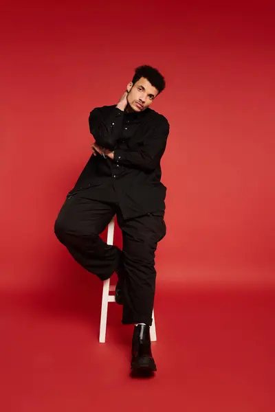A stylish young man sits on a stool, striking a confident pose against a bright red backdrop. — Stock Photo