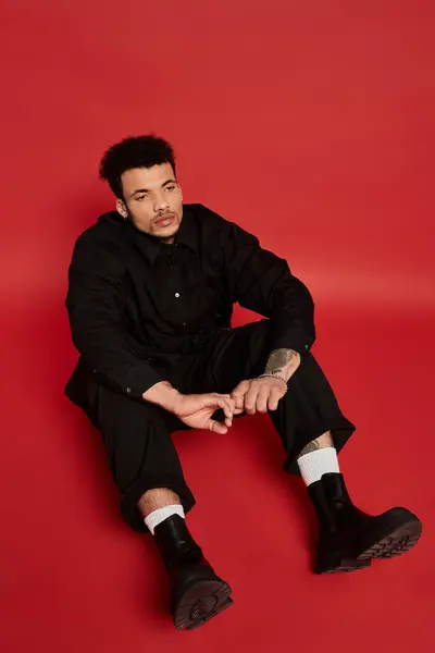 A young man with striking features sits casually on the floor, dressed in a black outfit. — Stock Photo