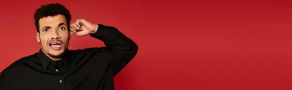 Handsome young man with curly hair shows surprise against a bold red backdrop wearing a black shirt — Stock Photo
