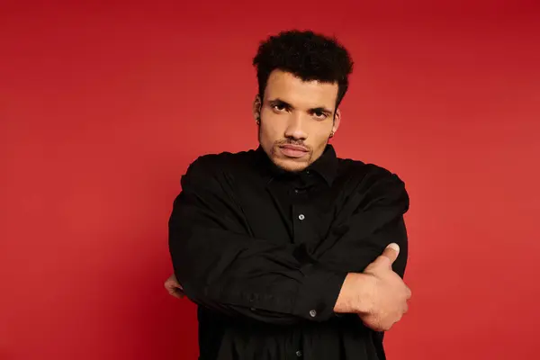 A young, handsome man with curly hair stands confidently with crossed arms before a red backdrop. — Stock Photo
