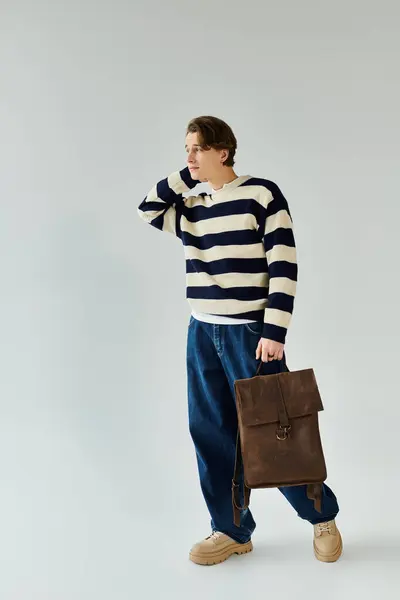 A handsome young man stands in a studio, dressed in a striped sweater and holding a backpack. — Stock Photo