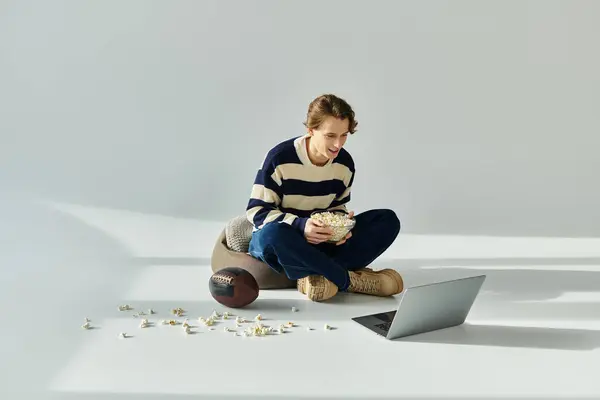 A youthful man sits cross-legged, delightedly munching popcorn while watching sports on his laptop. — Stock Photo