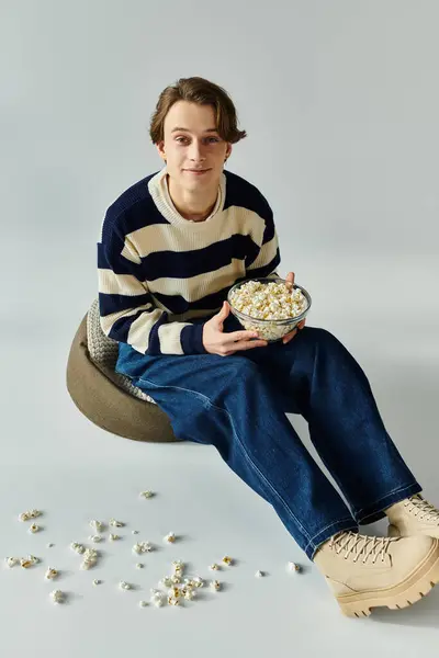 Bonito jovem em uma camisola listrada gosta de pipocas enquanto sentado em um estúdio elegante. — Fotografia de Stock