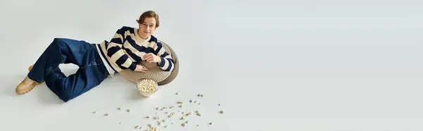 Um jovem bonito relaxa em uma superfície macia enquanto lanche pipocas em um estúdio, banner — Fotografia de Stock