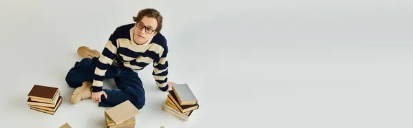 A young man in a striped sweater sits among books, exuding a casual yet studious vibe, banner — Stock Photo