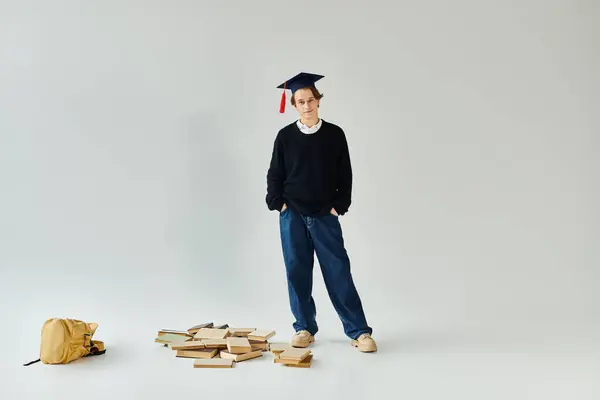 A young man proudly wears his graduation cap in a studio, surrounded by books and a backpack. — Stock Photo