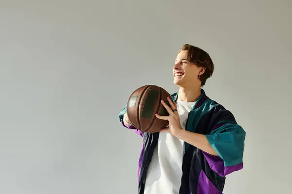 A young man wearing a fashionable bomber jacket joyfully holds a basketball in a studio. — Stock Photo