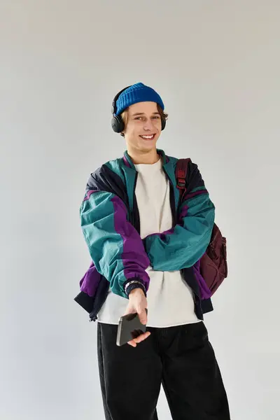 A young man wearing a colorful bomber jacket smiles while holding his smartphone in a studio. — Stock Photo