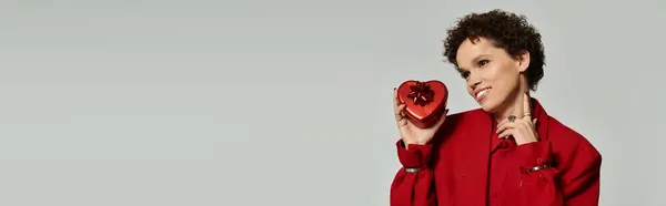 Une jeune femme en tenue rouge pose élégamment avec un cadeau en forme de cœur et un sourire ludique. — Photo de stock