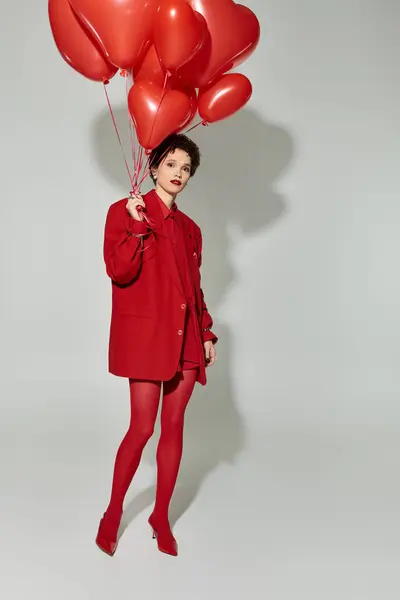 A stylish young woman dressed in red holds vibrant heart shaped balloons, exuding charm. — Stock Photo