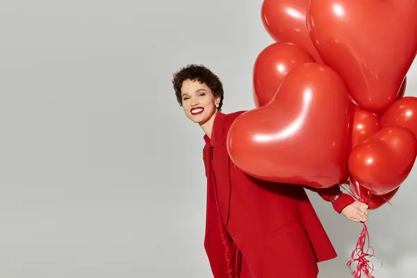 Eine junge Frau in auffallend rotem Gewand lächelt breit, während sie verspielt herzförmige Luftballons in die Höhe hält. — Stockfoto