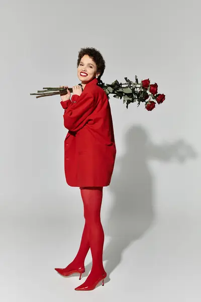 A joyful young woman radiates charm, dressed in vibrant red while playfully holding roses. — Stock Photo