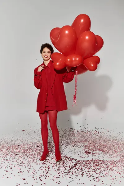 Une femme joyeuse en rouge tient des ballons en forme de cœur entourés de confettis colorés. — Photo de stock