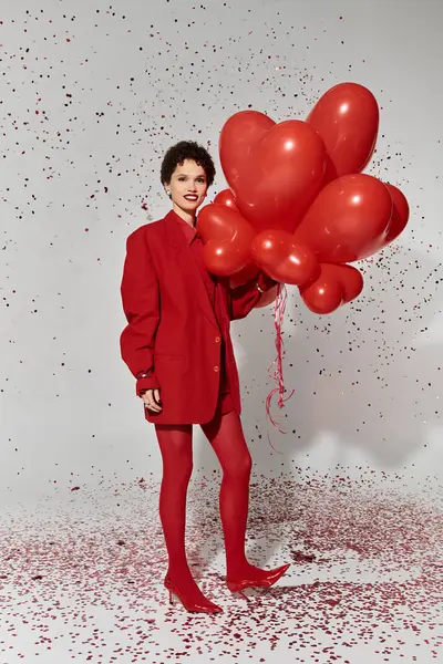 Une femme élégante en rouge tient joyeusement des ballons en forme de cœur au milieu des confettis. — Photo de stock