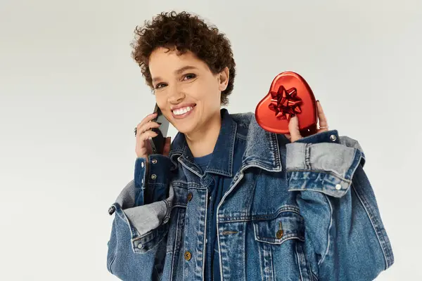 A joyful young woman with curly hair holds a heart shaped gift while chatting on her phone. — Stock Photo