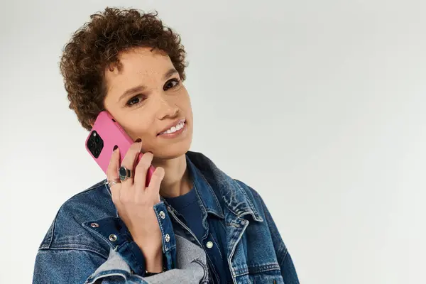 A young beautiful woman smiles while talking on her smartphone, wearing a denim jacket. — Stock Photo