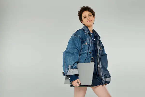 Confident young woman in oversized denim jacket poses with a laptop in a contemporary studio space. — Stock Photo