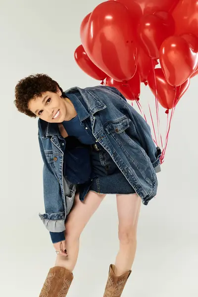 Une jeune femme joyeuse en denim pose avec des ballons rouges dans un studio lumineux. — Photo de stock