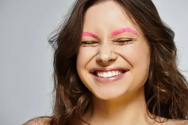 Young woman smiles confidently, showcasing her vibrant pink eyebrows against a grey backdrop. — Stock Photo
