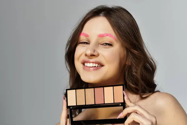 Une femme aux sourcils roses sourit tout en tenant des produits de maquillage sur fond gris. — Photo de stock