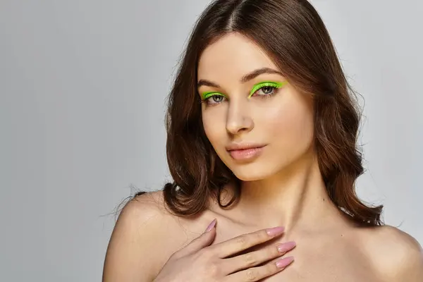 Mujer joven muestra sombra de ojos verde vibrante y maquillaje impecable contra un telón de fondo gris. — Stock Photo
