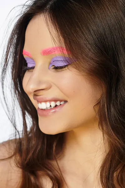 A young woman showcases her artistic makeup with purple eyeshadow and pink brows on grey. — Stock Photo