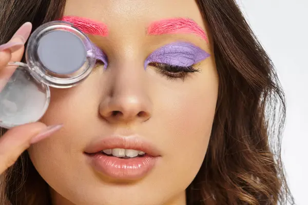 A young woman displays bold makeup with purple eyeshadow and pink eyebrows in a studio. — Stock Photo