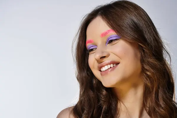 Una joven mujer exhibe vibrante sombra de ojos púrpura y cejas rosadas sobre un fondo gris. - foto de stock