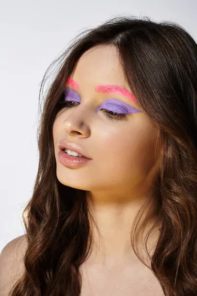 Pretty young woman showcases striking purple eyeshadow and pink eyebrows in a grey studio. — Stock Photo